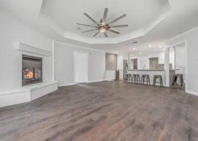 Bright, open living room and kitchen with dark wood flooring, white painted brick fireplace with fire burning, coffered ceiling with large modern ceiling fan, breakfast bar with metal stools and stainless steel appliances.