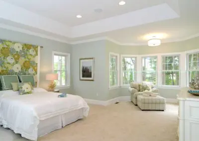 Light olive-green bedroom with a white bed and floral headboard, bedside table and lamp, bay windows overlooking trees, and a striped chaise lounge with ottoman