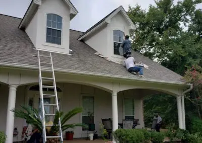 Workers repairing a suburban home's front porch and dormer windows — two men on the roof near a ladder working on a window, another on the porch, columns and lawn visible