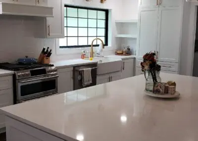 A bright, modern white kitchen with a large marble island in the foreground, farmhouse sink and gold faucet beneath a black-framed window, stainless steel range and dishwasher, white shaker cabinets with brass handles, and a small vase of flowers and decorative tray on the island.