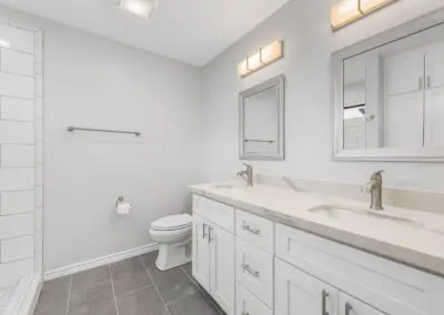 Bright, modern bathroom with double-sink white vanity, marble countertop, two mirrors and wall lights, tile shower on left and toilet near center, gray floor tiles.