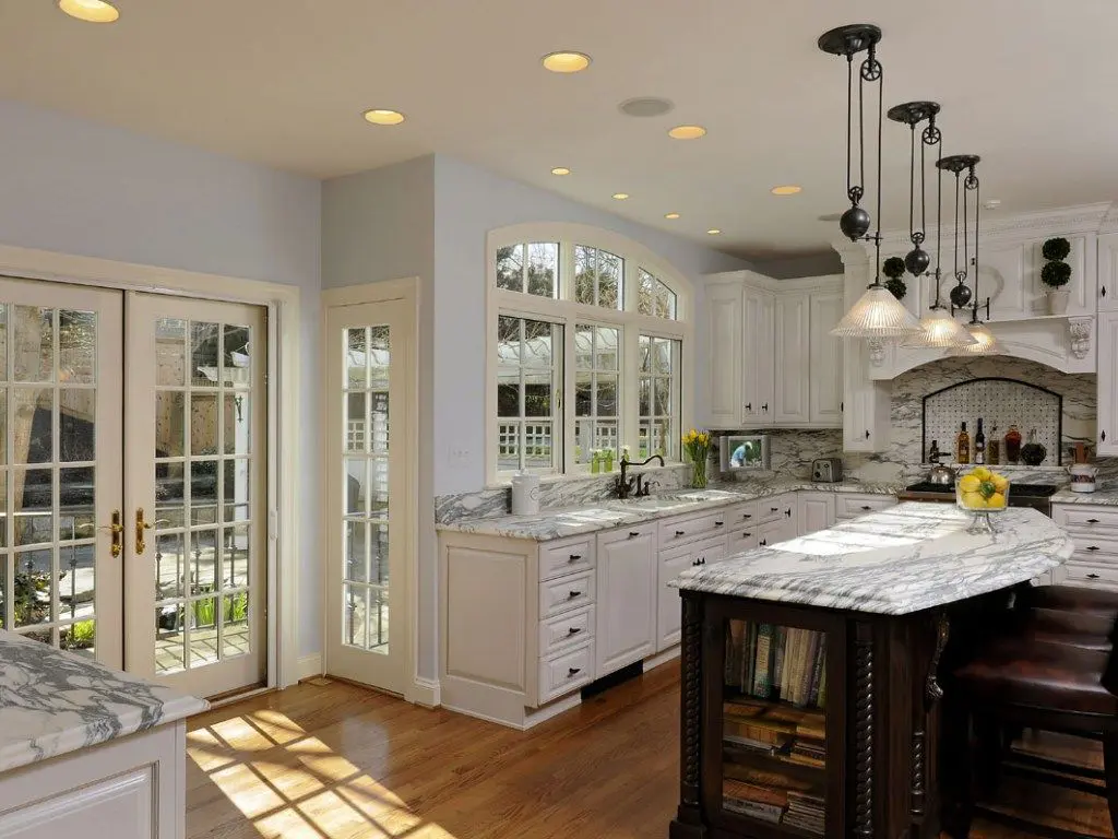 Bright, remodeled Bryan, TX kitchen with white cabinetry, marble countertops on island and perimeter, pendant and recessed lighting, hardwood floors, French doors to patio, large arched window over sink, and decorative range hood