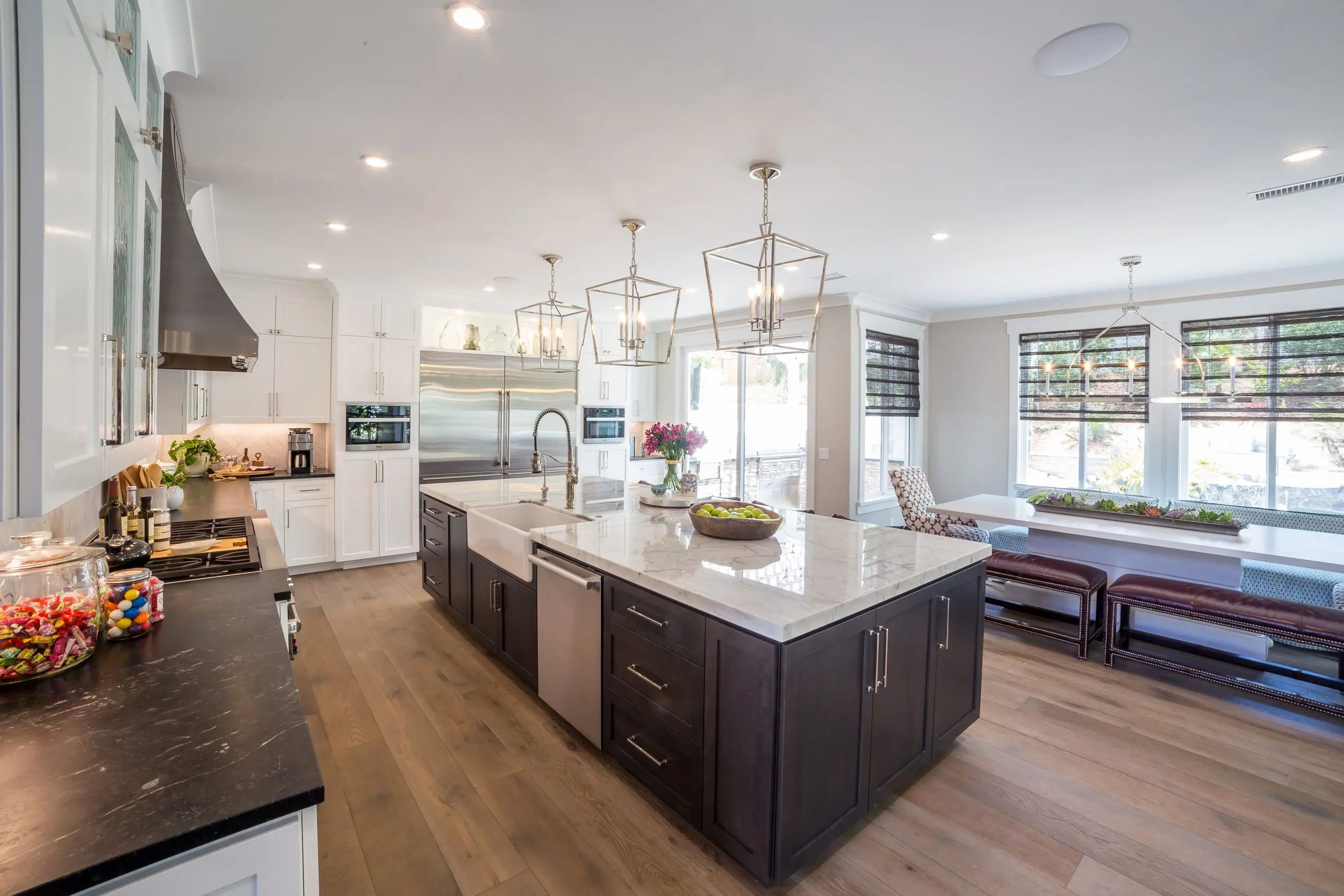 Spacious remodeled kitchen with large marble-topped island, dark lower cabinets and white upper cabinets, stainless steel appliances, farmhouse sink, pendant lighting, built-in bench seating by bright windows — modern home remodeling and kitchen renovation showcase.