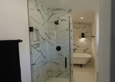 Bathroom remodel showing a glass-enclosed shower with black fixtures and white marble-look tile, a freestanding clawfoot tub in the background, and a black towel on a wall-mounted bar