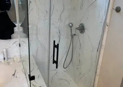 Glass-enclosed bathroom shower with white marble-patterned wall and bench, overhead rain showerhead, handheld shower wand, black metal door handle and hinges, and tiled floor visible alongside a bathtub edge