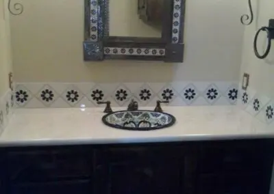Bathroom vanity with dark wood cabinet, decorative tile backsplash with blue floral motif, patterned ceramic sink inset in light countertop, ornate framed mirror centered above, and wall sconces on either side