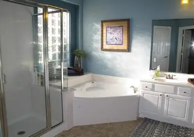 Bright remodeled bathroom with corner jetted soaking tub, glass-enclosed shower with chrome frame, white vanity and sink, blue painted walls, framed wall art, and patterned floor mat