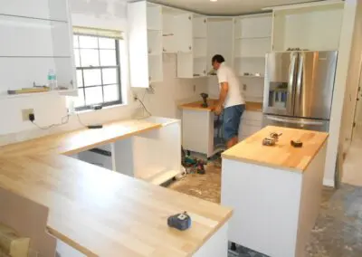 Worker installing white kitchen cabinets and butcher-block countertops during a cabinet installation in a residential kitchen, tools and refrigerator visible