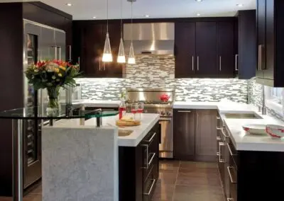 Modern kitchen renovation with dark wood cabinets, white quartz countertops, mosaic tile backsplash, stainless steel refrigerator and range, pendant lights over island with glass breakfast bar and vase of flowers
