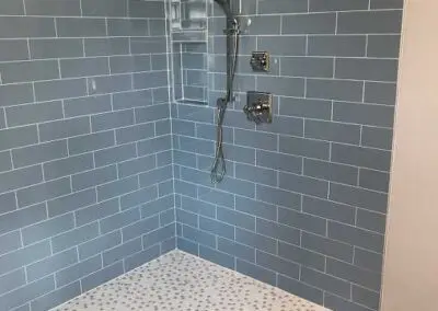 Shower with light blue subway tile walls, recessed niche with glass shelf, chrome handheld showerhead and controls, white pebble-pattern floor tile and small frosted window at top left
