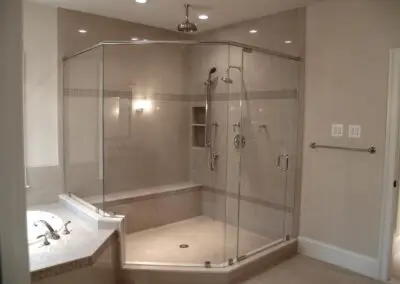Walk-in glass shower enclosure with tiled bench, overhead rain shower and hand shower, chrome fixtures, and adjoining tiled bathtub in a neutral beige bathroom